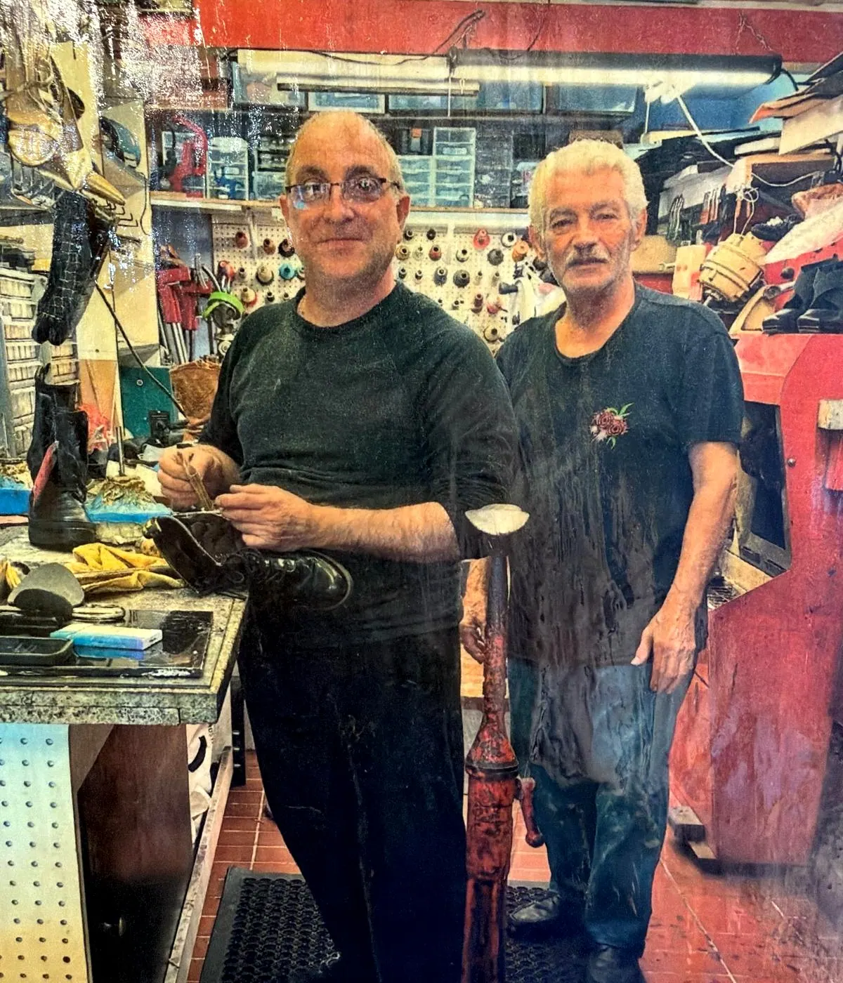 Two of the team at the workbench inside Robert's Shoe Repair, one holding a boot he is working on, with lasts, tools and finished shoes around them