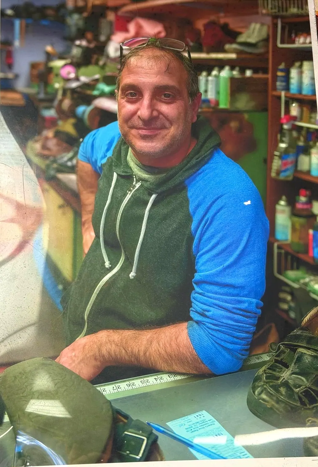 One of the team at the workbench at Robert's Shoe Repair, a shoe and a measuring rule on the counter in front of him and shelves of leather care behind